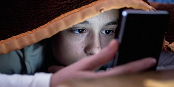 Menino com aproximadamente 10 anos, embaixo do cobertor, conferindo o celular, no escuro