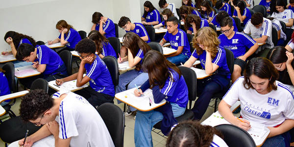 Alunos do Colégio Classe A estão sentados nas carteiras realizando provas de Olimpíadas do Conhecimento. 