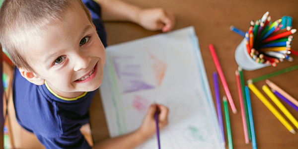 menino com cerca de 3 anos sorrindo, com lápis de cor e seu desenho 